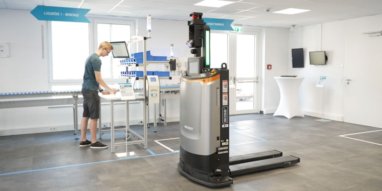 Automatisierter Stapler (FTS) transportiert Waren direkt vom Lager zur Montagelinie, wo sie vom Werker entgegengenommen werden, als Beispiel für sicheren Einsatz von FTS in Lager und Produktion.